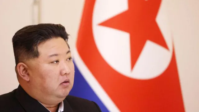 North Korean leader Kim Jong Un looking to the side. Behind him is a large North Korea flag.