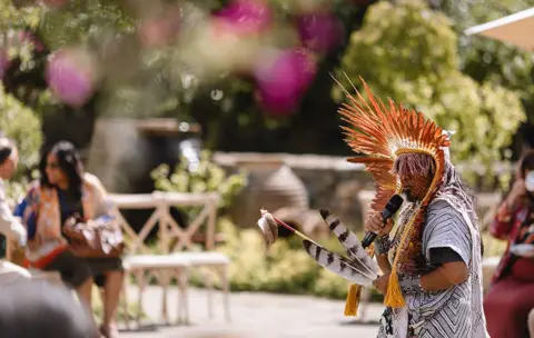 A man wearing traditional dress holds two black and white feathers and a microphone at Highgrove.