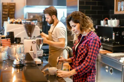 A young woman with long wavy hair and a red checked shirt is decorating a cup of cappuccino. A young bearded male barista, wearing a beige apron and grey t-shirt, is working in the background amidst steam rising from an espresso machine. The café has a sleek black-tiled wall and modern coffee-making equipment, creating a warm and inviting workspace atmosphere.