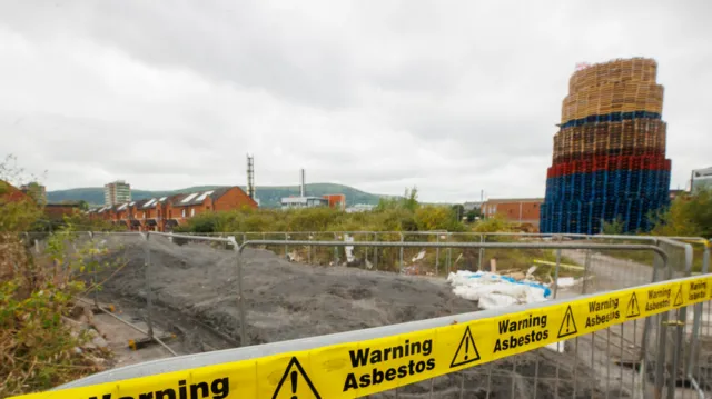 Yellow police tape warning of asbestos danger in front of a tall bonfire, with houses in the background.