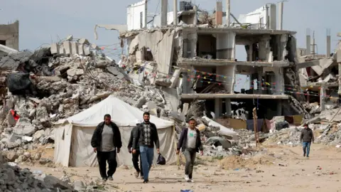 Palestinians walk among rubble in Rafah, with a tent and a bombed out building in the background