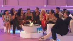 ITV/Shutterstock Islanders gathered around the fire pit in the Love Island villa. 13 islanders sit tightly on the white L-shape sofa, dressed smartly and holding drinks. The villa is lit with pink and blue lights.