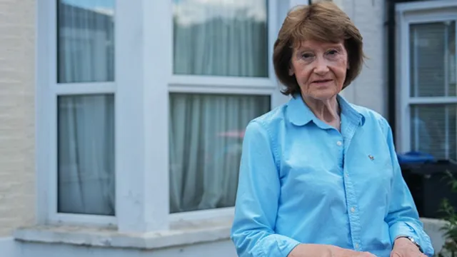 Sue stands in front of a light-coloured brick or stone terraced house. She is wearing a light blue button-up shirt with sleeves rolled up to the elbows and a small embroidered logo on the left chest. Behind her, the house features two large white-framed windows with sheer curtains and a ledge beneath each window. The overall scene is well-lit, and no additional objects or textures are visible