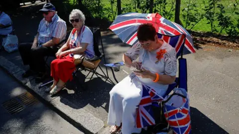 A spectator shading herself from the sun with a union jack umbrella. She is sitting down and wearing a while skirt and white top, she has short red-brown hair. To her right are a couple sitting on folding chairs. The woman is in a red dress and the man is in a striped red-white-and-blue shirt and dark blue cap.
