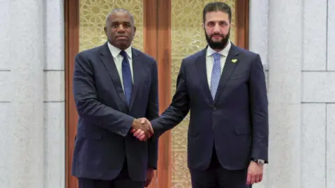 Ahmad al-Sharaa and David Lammy shake hands, both in suits and looking towards the camera.