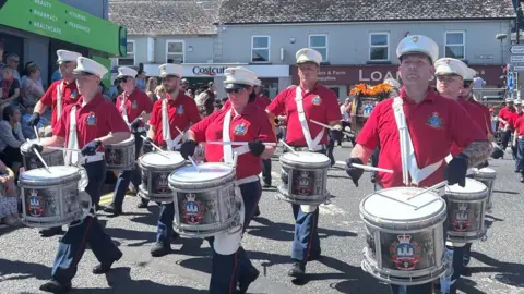 A parade of band members marching through Kesh wearing red short sleeved shirts, white hats and playing white drums. The sun is shining on the band members.