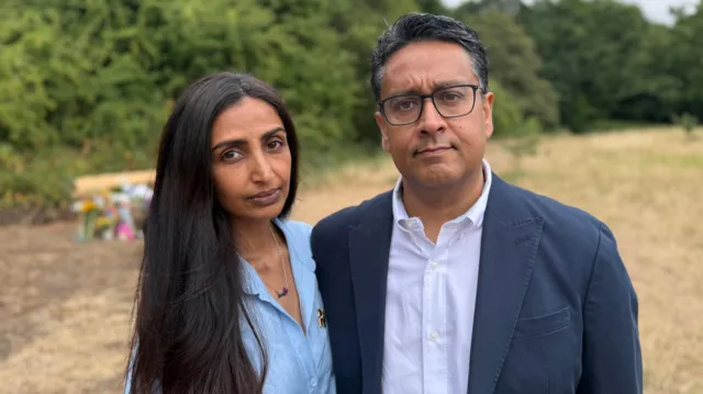 Smera Chohan and Sajjad Butt, Nuria's parents, look towards the camera while standing in front of a wooden bench commemorating their daughter Nuria.