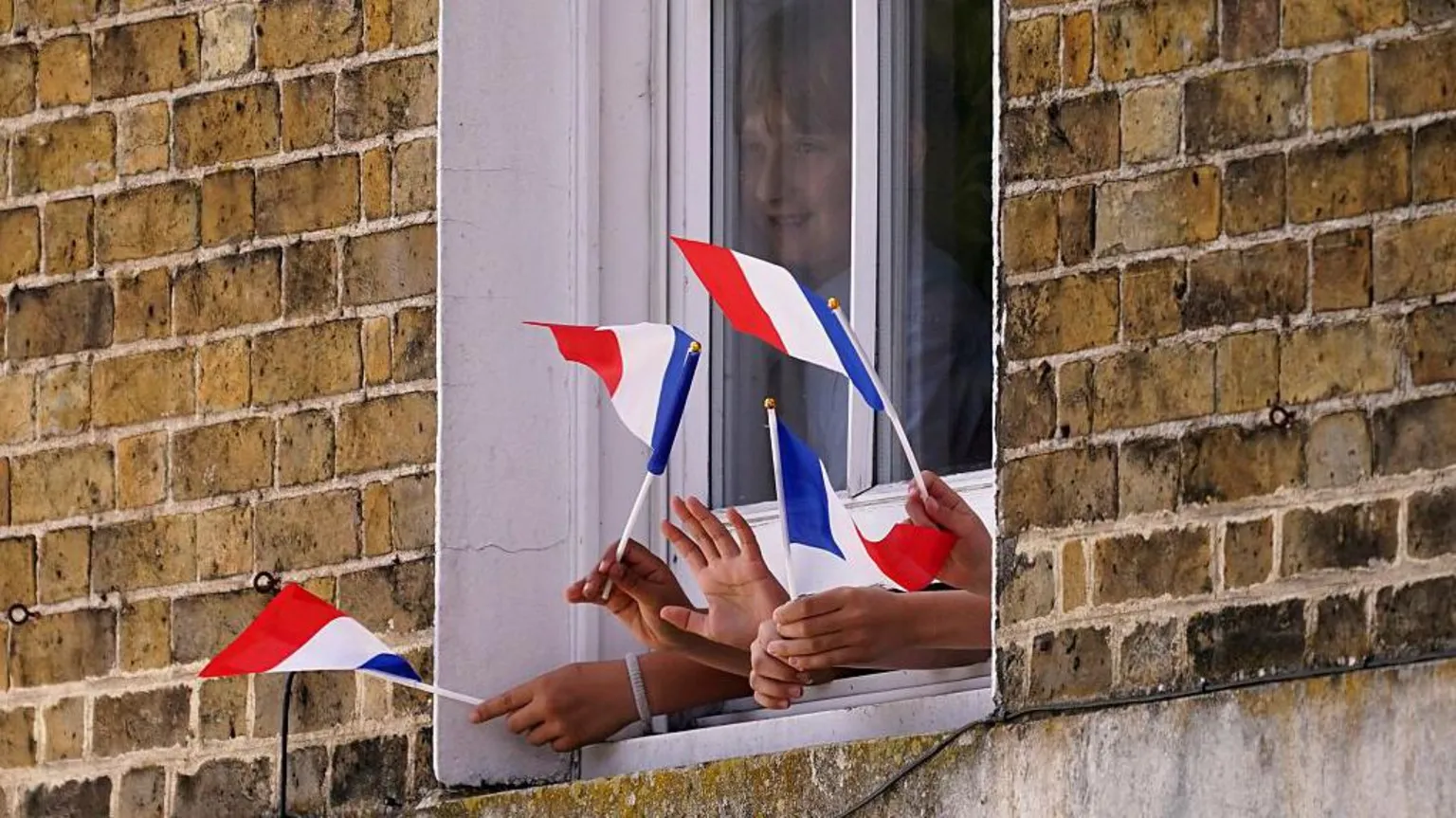 AFP via Getty Images A young boy is seen behind a window which has his hands and the hands of other young people sticking out of a gap at the bottom of it while waving the red, white and blue flag of France.
