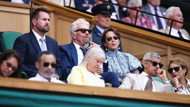 Richard Branson sits in the Royal Box on Centre Court alongside a woman wearing a blue dress and a man wearing a blue suit.