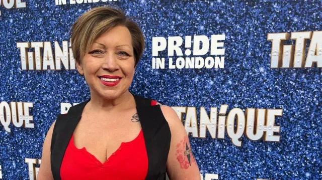 Dee Llewellyn, smiling at the camera in front of a Pride in London branding board.