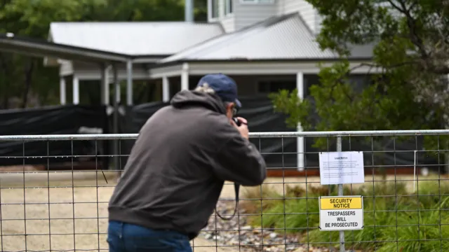EPA A photographer takes a picture over the gate looking towards a white house with black tarpaulin over the carport and windows. A sign saying security notice, trespassers will be prosecuted hangs on the metal gate. There are trees and grass just beyond the gate.