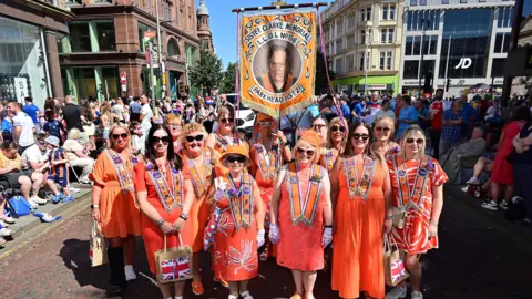 A group of women in orange dresses and sashes wearing sunglasses as they pose on a street in Belfast and hundreds of spectators are behind them. The women are holding a banner that says "Sister Clarke Memorial L.L.O.L No. 156 Parkhead Dist 2". The sun is shining on the women.