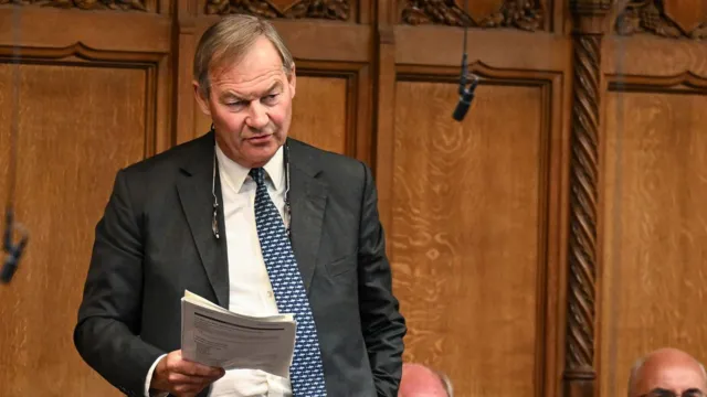 PA Media Rupert Lowe stands in the House of Commons, holding a document as he speaks. He is surrounded by seated members of Parliament. The chamber features traditional green benches and ornate wooden paneling.