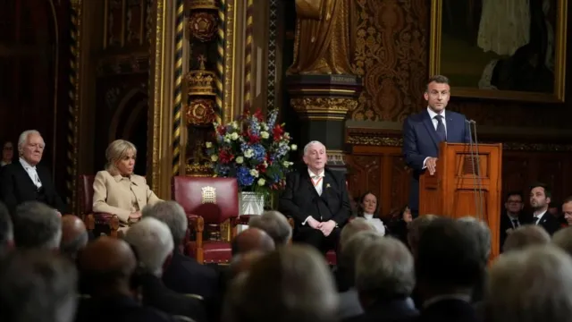 Macron stands at a lectern inside Parliament. Behind him, Speaker Lindsay Hoyle sits with his hands folded in his land, and to his left sits Brigitte Macron