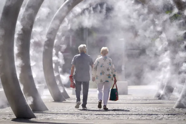 A man and a woman are walking inside ring metal structures that are spraying mist on a sunny day.