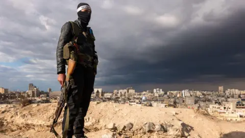 A fighter with Hayat Tahrir al-Sham, or HTS, pauses with a Kalashnikov rifle while standing above the city of Homs.