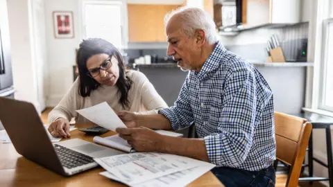 Older man and woman sitting at a kitchen table with paperwork and a laptop in front of them.