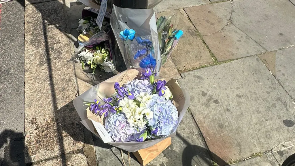 Bouquets of flowers have been placed on a pavement near where the stabbing occurred, with police tape visible.
