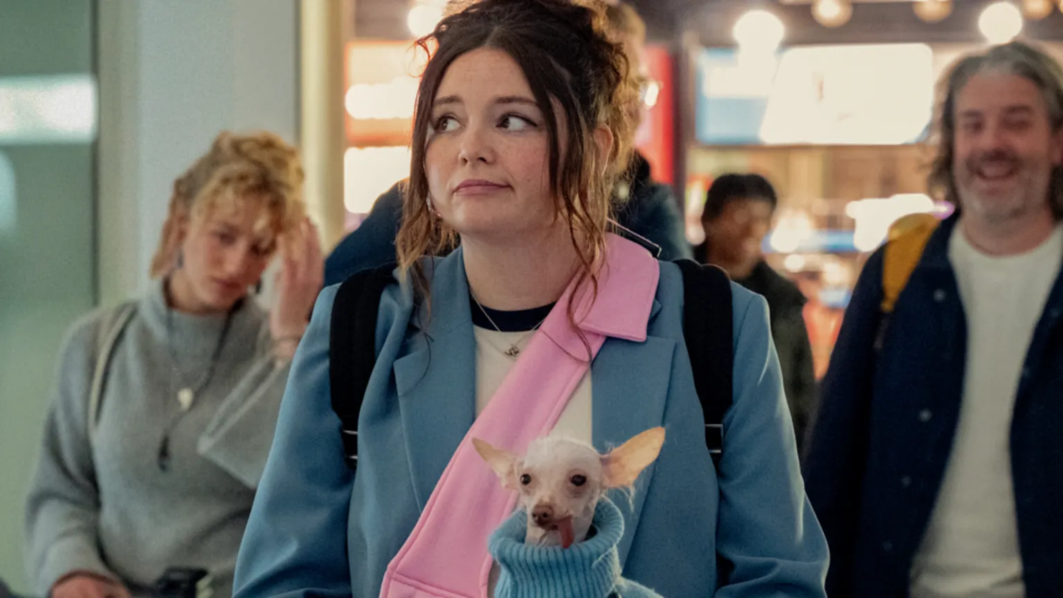 Megan Stalter as Jessica in Too Much, wearing a blue coat and a pink bag, carrying a small dog in a blue jumper