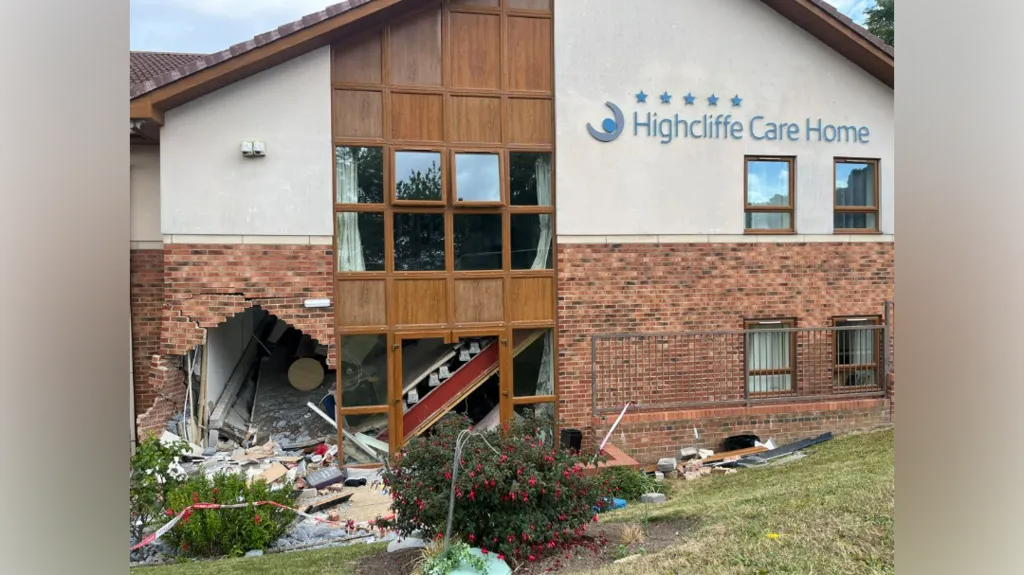 A brick and stone building with the sign reading \'Highcliffe Care Home\' in blue writing. A large hole can be seen at the bottom left of the building next to a window. Bricks and debris from the wall are scattered all around and in the bushes and grass in front of the building.