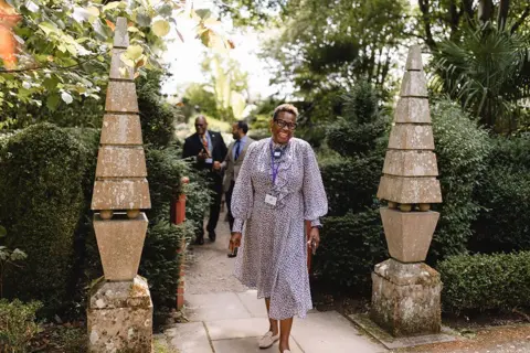 A woman wearing a blue and white dotted dress smiles as she walks between two stone pillars at Highgrove.
