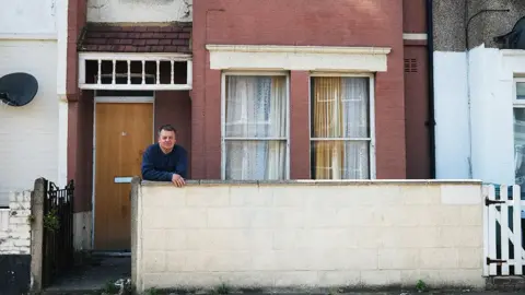 Roye Chapman is leaning on a low wall in front of a red brick house with two windows covered by lace curtains and a wooden door. The house has a small front yard with some grass and plants. Roye is wearing a blue long-sleeve shirt