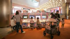 Backview of children and parents sitting and standing in front of colourful arcade machines in a brightly lit mall.