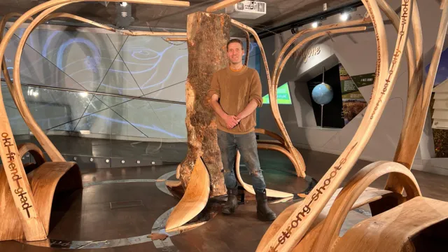 Artist Charlie Whinney stands beside his sculpture, which incorporates the Sycamore Gap tree trunk and curved wooden elements forming benches.